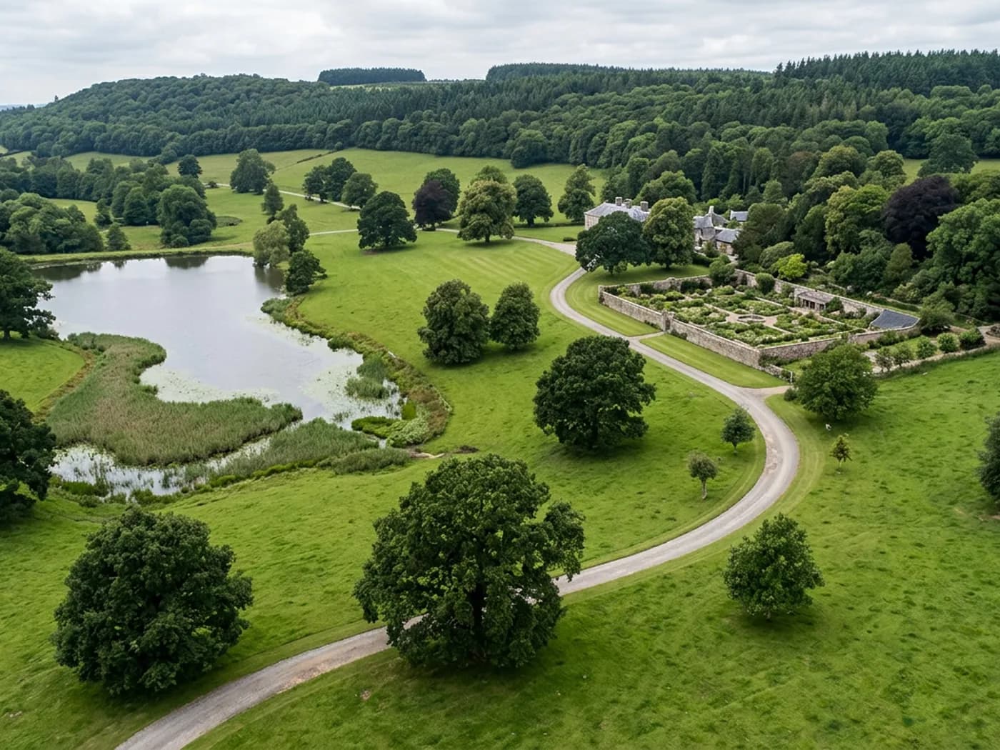 Parkland on a Devon estate from the air, mature oak and beech standing in cropped grass with a lake, a gravel drive and a walled garden